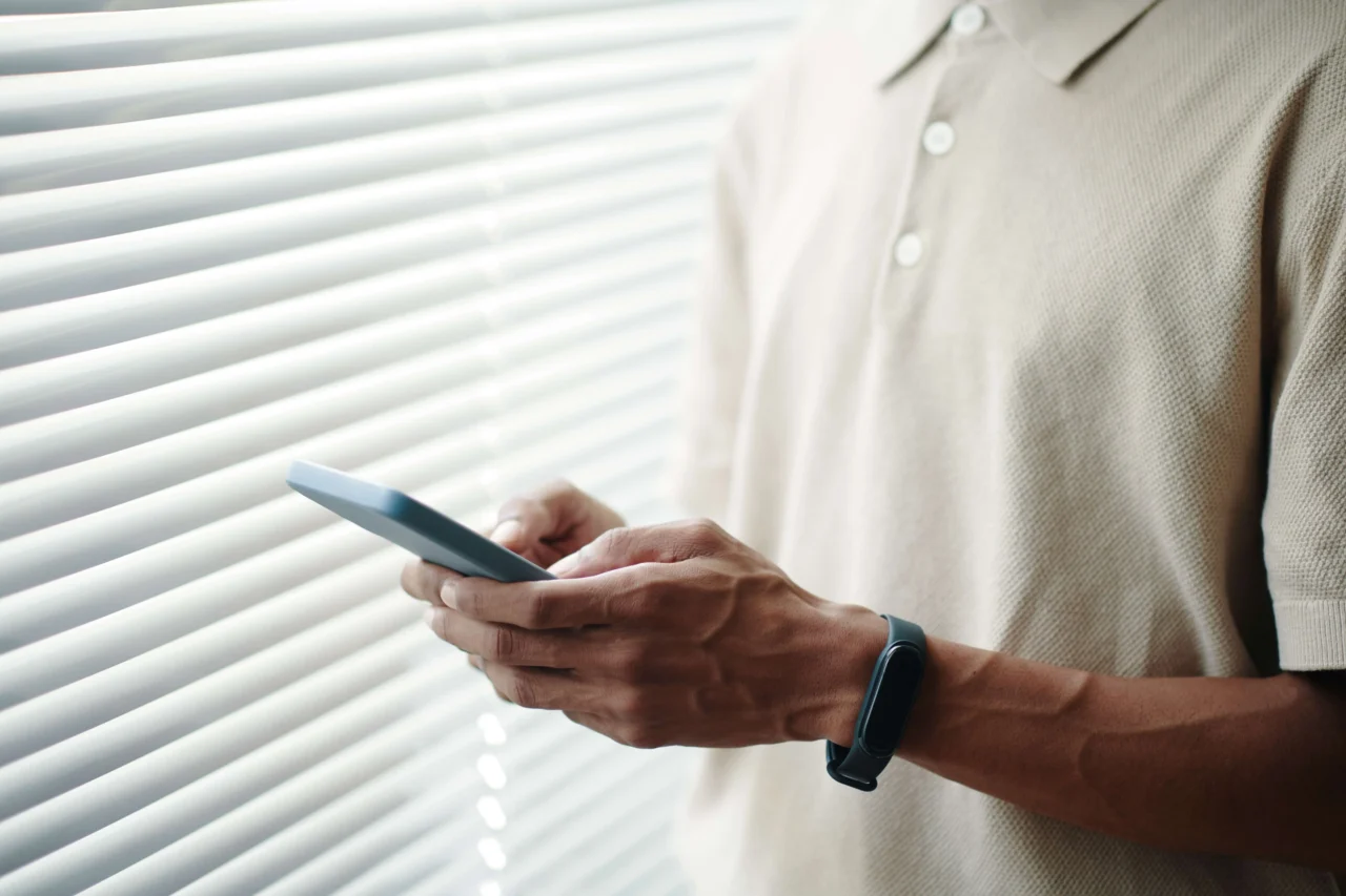 Hombre usando el móvil junto a una ventana con persianas, revisando aplicaciones de pago en línea.